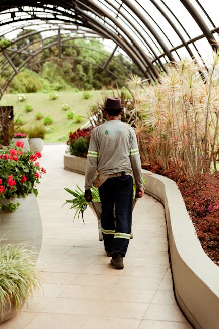 A gardener walking along a curved pathway inside a greenhouse, carrying a small bunch of green plants in one hand while wearing a grey uniform with reflective stripes, black gloves, and a wide-brimmed